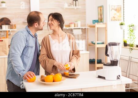 Frullato per la preparazione di coppia in cucina . Donna che si stacca di arancio mentre sorride al marito. Stile di vita sano e spensierato, dieta e preparazione della colazione in un'accogliente mattina di sole Foto Stock