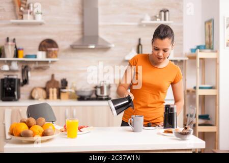 Donna che versa caffè caldo in tazza al mattino dalla pentola. Casalinga a casa, preparare caffè macinato fresco in cucina per colazione, bere, macinare caffè espresso prima di andare al lavoro Foto Stock