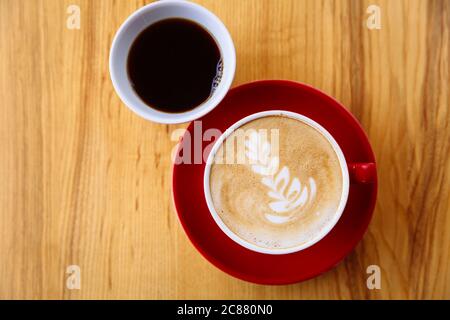 Caffè aromatico in una tazza rossa con schiuma di latte e latte art e caffè appena fatto in una tazza bianca su un tavolo di legno chiaro. Disposizione piatta Foto Stock