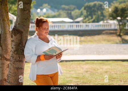 Tempo libero e istruzione. Una donna sovrappeso in un vestito che legge un libro in un parco, appoggiata su un tronco di albero. Spazio di copia. Foto Stock