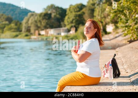 Una donna felice in sovrappeso si siede sul bordo del molo, stringendo un libro al suo petto. Sullo sfondo, il mare e il Parco. Foto Stock