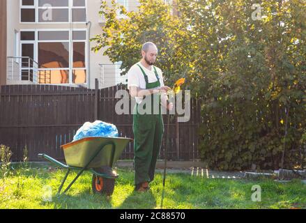 Il giardiniere stanco in uniforme verde pulisce il rastrello dalle foglie. Giardinaggio e pulizia del cortile. Foto Stock