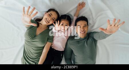 Madre cinese e padre che giace con figlia carina sul letto guardando la macchina fotografica con le braccia sollevate Foto Stock