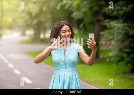 Allegra donna nera che comunica online con la famiglia o gli amici su smartphone al parco Foto Stock