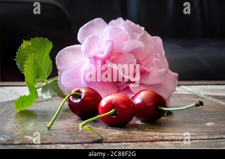 Primo piano di tre ciliegie rosse con rosa e foglie verdi su una superficie marrone. Macro, sfondo scuro, illustrazione Foto Stock