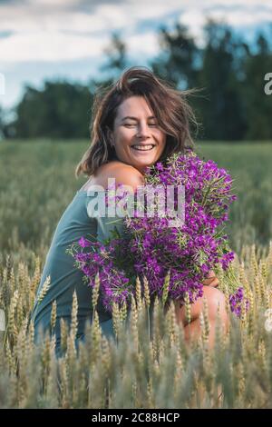 Bella donna in un vestito blu carino e un enorme bouquet di fiori viola, ride e si siede su campo all'aperto in un prato durante il tramonto. Livesty Foto Stock