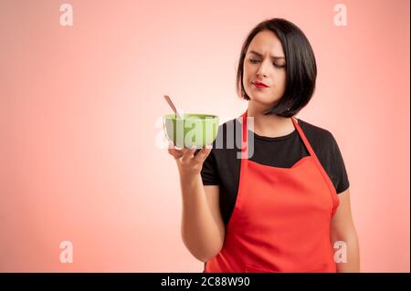 Donna impiegata al supermercato con grembiule rosso e t-shirt nera, tenendo e guardando la ciotola in mano isolata su sfondo rosso Foto Stock