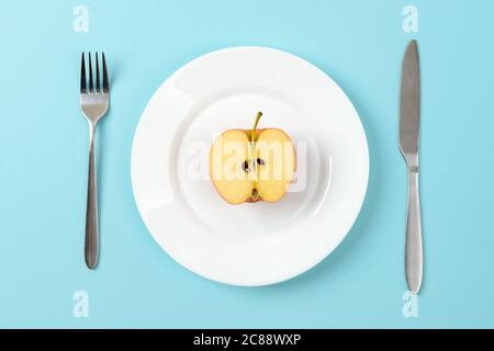 Metà di mela matura succosa su un piatto da pranzo bianco, coltello da tavola e forchetta su sfondo blu. Concetto di alimentazione sana, vegetarianismo, dieta di cibo crudo. Foto Stock