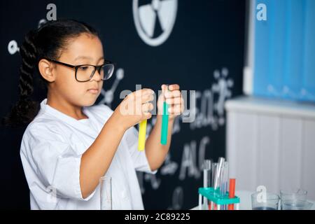 Cute pupilla vietnamita femminile in occhiali protettivi e uniforme bianca, facendo ricerca biochimica in classe chimica Foto Stock