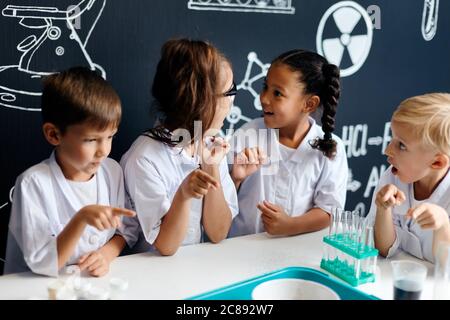 Gruppo di bambini multirazziali diversi in abiti bianchi scienziati facendo esperimenti scientifici con reagenti in laboratorio, essendo sorpreso e stupito Foto Stock