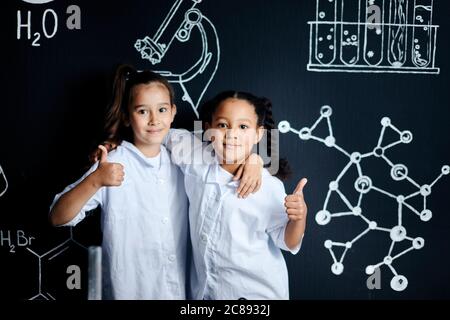 Due ragazze in abiti bianchi, pochi futuri scienziati, guardando la macchina fotografica mentre si visita la scuola labaratory, in piedi con pollici contro la lavagna nera Foto Stock