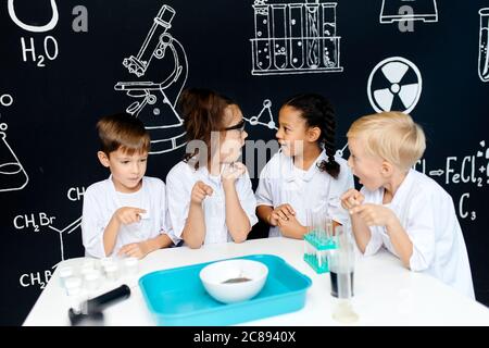 Gruppo di bambini multirazziali diversi in abiti bianchi scienziati facendo esperimenti scientifici con reagenti in laboratorio, essendo sorpreso e stupito Foto Stock