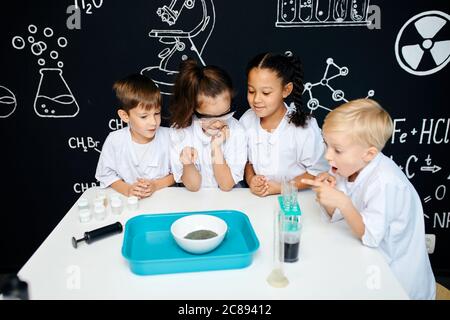 Gruppo di bambini multirazziali diversi in abiti bianchi scienziati facendo esperimenti scientifici con reagenti in laboratorio, essendo sorpreso e stupito Foto Stock