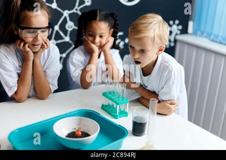 Gruppo di bambini multirazziali diversi in abiti bianchi scienziati facendo esperimenti scientifici con reagenti in laboratorio, essendo sorpreso e stupito Foto Stock