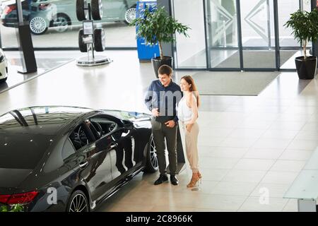 Di acquistare la loro prima vettura insieme. Angolo di Alta Vista del giovane venditore auto in piedi presso la concessionaria dicendo circa le caratteristiche della vettura di committenti. Foto Stock