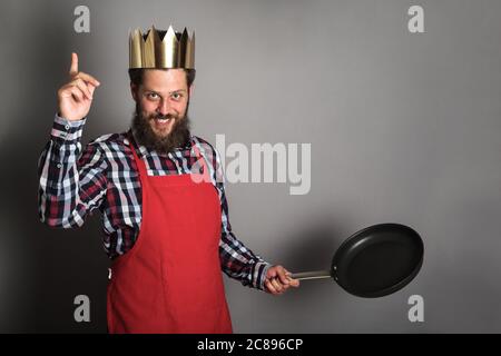 Re di cucinare con il dito in su, divertente uomo bearded in corona di carta e cuoco grembiule con padella vuota è pronto a fare una lezione Foto Stock