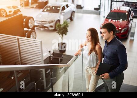 Acquistare la prima auto insieme. Vista ad alto angolo di un giovane venditore di automobili al piano superiore presso la concessionaria che illustra le caratteristiche della vettura alla donna Foto Stock