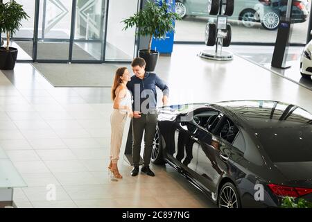 Di acquistare la loro prima vettura insieme. Angolo di Alta Vista del giovane venditore auto in piedi presso la concessionaria dicendo circa le caratteristiche della vettura di committenti. Foto Stock