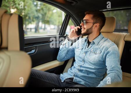 sorridente, un ragazzo fantastico sta parlando al telefono mentre si è seduti in auto. primo piano vista laterale foto. conversazione, concetto di tecnologia Foto Stock