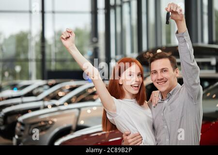 Concetto di concessionaria di auto in visita. La coppia con capelli rossi eccitati e sorridi festeggia l'acquisto di una nuova auto, tenendo la chiave in mano, guardando la fotocamera e sorridendo. Foto Stock