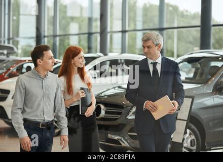 Business auto, concessionaria auto, clienti e concetto di Rep. Venditore maturo che parla con una giovane coppia nello showroom della concessionaria. Foto Stock