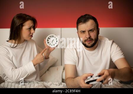 la ragazza punta a orologio mentre il suo ragazzo gioca i videogiochi a letto di notte Foto Stock