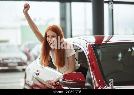 La giovane donna driver dai capelli rossi che gridava di gioia e mostra la chiave di una nuova auto mentre si siede in un'auto che lei riceve come regalo per il b-day. Foto Stock
