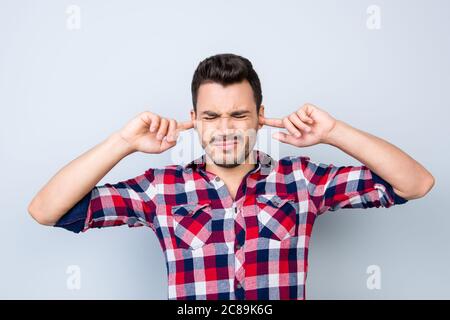 Basta! Giovane hipster ragazzo brunet sta coprendo le orecchie a causa di rumori, è stressato e stanco, in camicia a scacchi su sfondo chiaro Foto Stock