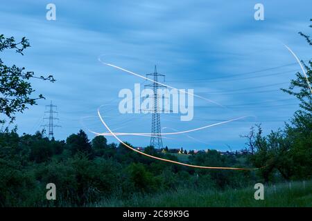 Grande elettricità pilone anche linea aerea pilone si trova dietro prato verde albero, arancione effetti di luce può essere visto. Germania. Foto Stock