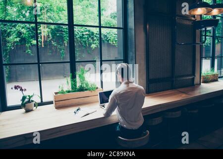 Vista posteriore di un giornalista elegante ragazzo scrivere una storia in un posto di lavoro in loft stile coworking, ben vestito, seduto vicino alla finestra con vista sul giardino Foto Stock