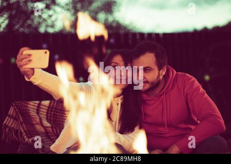 Seppia tonica ritratto di coppia amorevole seduta dal fuoco in natura che prende selfie Foto Stock