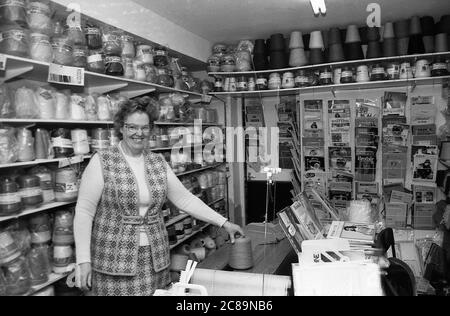 Anni 80, storico, proprietario femminile di un negozio di vendita di lana in piedi nel suo negozio, mostrando la vasta gamma di diversi lane disponibili e riviste di maglieria Inghilterra, Regno Unito. Foto Stock