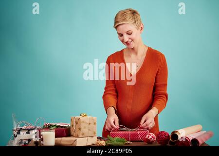 Sorridente saler incinta preparare regali per i clienti al suo negozio di articoli da regalo al tempo di natale facendo un arco sulla scatola regalo rossa, isolato in studio sopra la ba blu Foto Stock