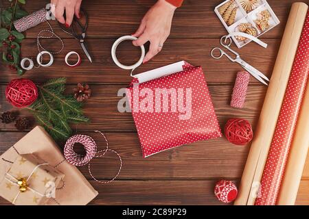 Colpo d'asta delle mani del progettista femminile che impacchettano i regali di Natale in fogli di imballaggio sulla tabella di legno con gli articoli di decorazione e i baubles di Natale Foto Stock