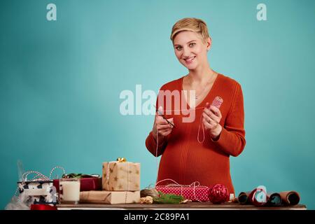 Bella bionda giovane madre imballaggio regali di Natale per i suoi figli azienda forbici e backer s striscia dello spago in mani, isolato in studio su blu Foto Stock