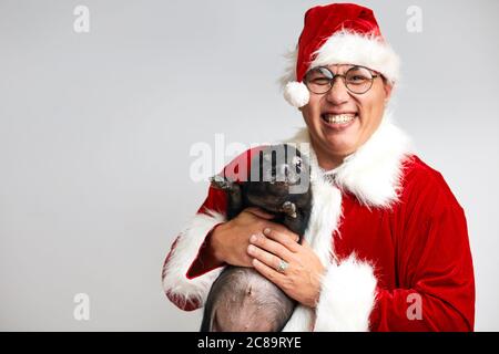 L'uomo asiatico felice in occhiali e Babbo Natale suit tiene nero piccolo piggy in mani sorridenti ampiamente alla macchina fotografica che desidera buon natale, isolato sopra Foto Stock