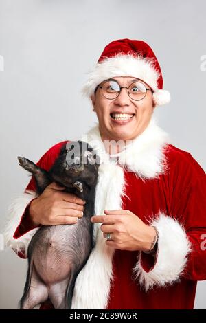 L'uomo asiatico felice in occhiali e Babbo Natale suit tiene nero piccolo piggy in mani sorridenti ampiamente alla macchina fotografica che desidera buon natale, isolato sopra Foto Stock