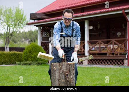 Uomo adulto in jeans camicia e tute tagliere di legno con ascia in campagna Foto Stock