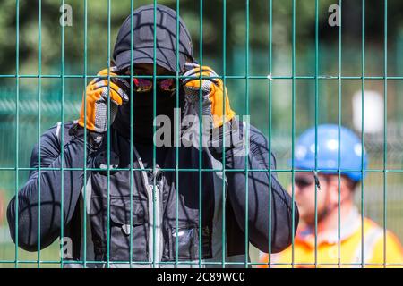 Harefield, Regno Unito. 22 luglio 2020. Una guardia di sicurezza mascherata HS2 osserva un fotografo della stampa durante i lavori per il collegamento ferroviario ad alta velocità HS2 vicino a Harvil Road. Gli attivisti ambientali della HS2 Rebellion e della Stop HS2 continuano a protestare contro la HS2, che attualmente è prevista per costare £106 miliardi e rimarrà un contributo netto alle emissioni di CO2 durante i suoi 120 anni di vita previsti, da una serie di campi di protezione della fauna selvatica lungo il suo percorso. Credit: Mark Kerrison/Alamy Live News Foto Stock