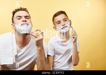 padre e suo figlio con la schiuma da barba sul viso si stanno radendo e guardando la macchina fotografica Foto Stock