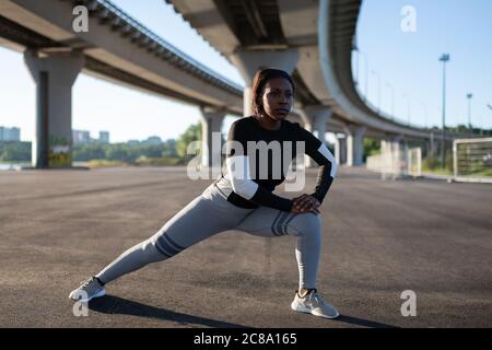 Corridore etnico che si affondo e allunga le gambe durante l'allenamento mattutino sulla strada della città Foto Stock