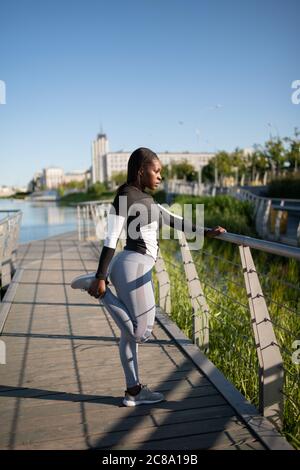 Vista laterale della gamba nera che stretching runner durante l'allenamento mattutino in banchina in città Foto Stock