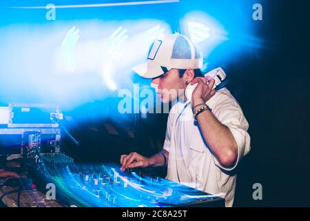DJ con cuffie durante la festa notturna sotto la luce blu e la gente si affollano in background Foto Stock