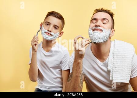 padre e suo figlio con la schiuma da barba sul viso si stanno radendo e guardando la macchina fotografica Foto Stock