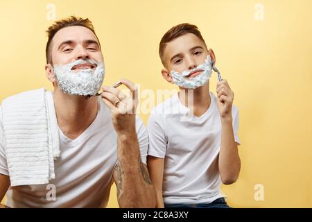 padre e suo figlio con la schiuma da barba sul viso si stanno radendo e guardando la macchina fotografica Foto Stock