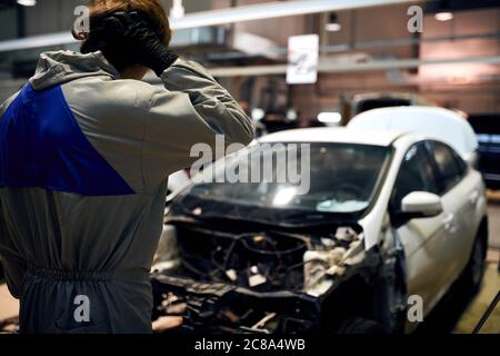 uomo meccanico stressato in bianco uniforme che tiene testa contro l'auto in garage di riparazione Foto Stock