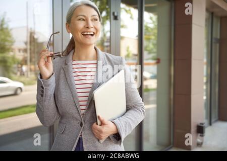 Pronto al lavoro. Bella e felice donna d'affari matura che tiene il laptop e sorride mentre si trova vicino a un edificio d'ufficio. Uomini d'affari e tecnologie moderne, successo, concetto di lavoro Foto Stock