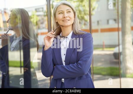 Donna di successo. Ritratto di una bella donna d'affari felice matura in classico indossare occhiali, guardando da parte e sorridendo mentre si sta in piedi all'aperto. Uomini d'affari Foto Stock