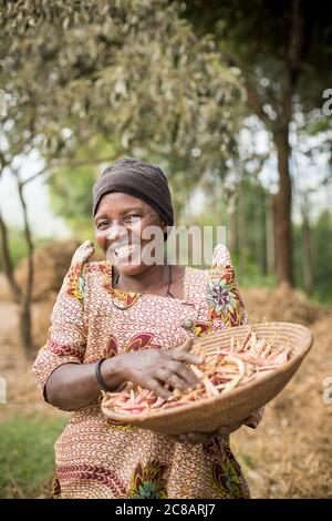 Una piccola contadina sorridente tiene un cesto della sua coltura di fagioli appena raccolta nella sua fattoria nel distretto rurale di Lyantonde, Uganda, Africa orientale. Foto Stock