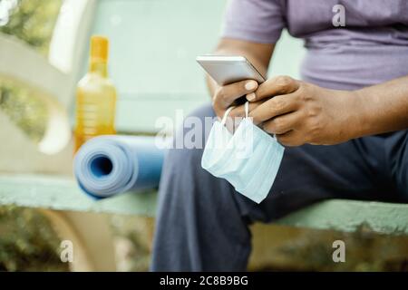 Uomo che usa il cellulare con maschera in mano al parco durante lo yoga di mattina o allenamento con tappetino - concetto di coronavirus o covid-19 consiglio, protezione o a. Foto Stock